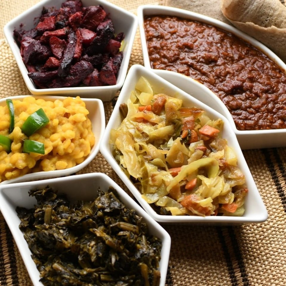 Five white bowls filled with various colorful Ethiopian dishes, including injera bread in the background.